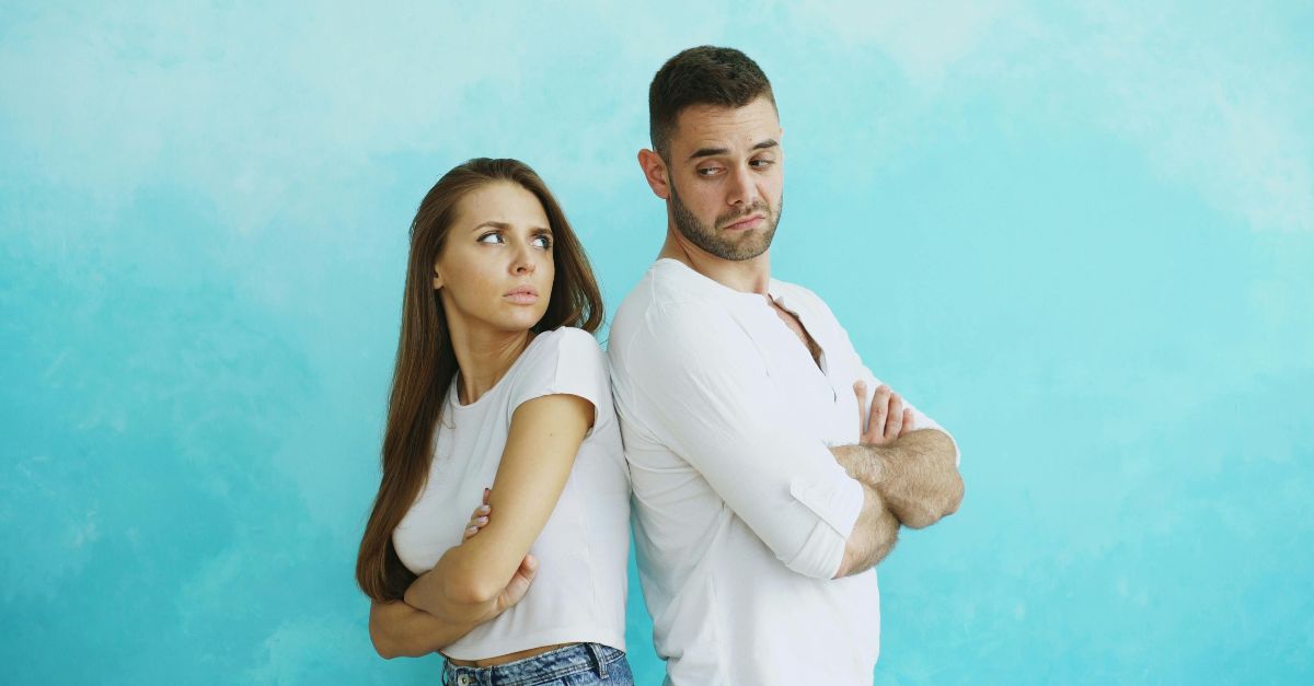 Young couple stands back to back with crossed arms against blue background.