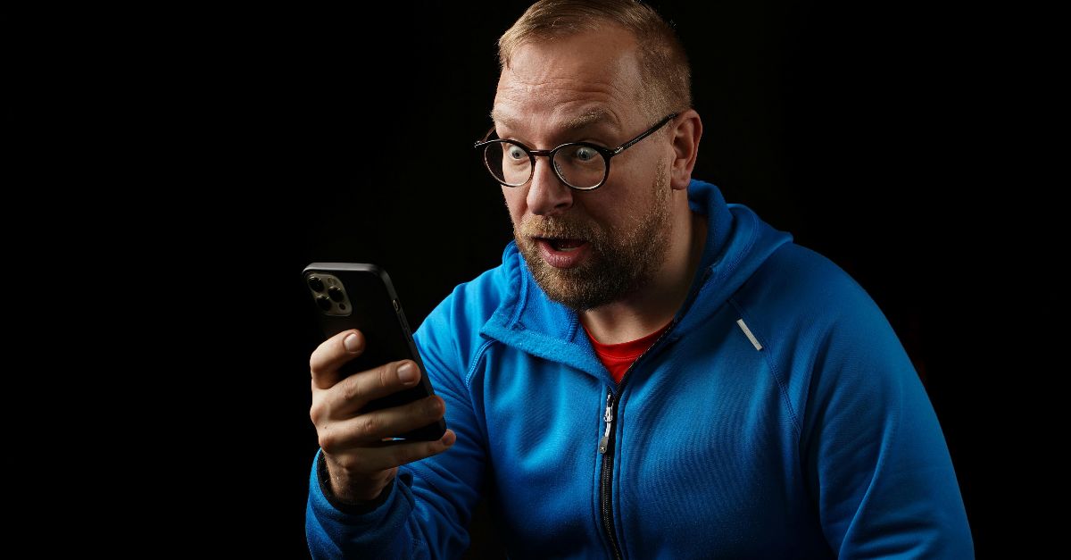 Surprised man in blue jacket looking at smartphone against a black background.