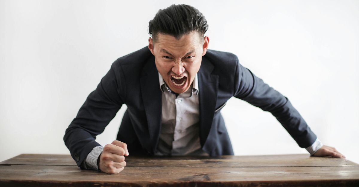 Expressive angry businessman in formal suit looking at camera and screaming with madness while hitting desk with fist