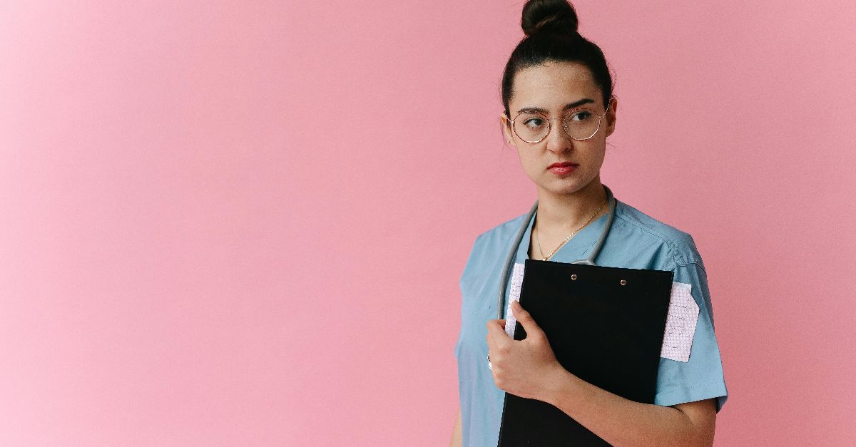 Confident female doctor in scrubs holding a clipboard against a pink background.