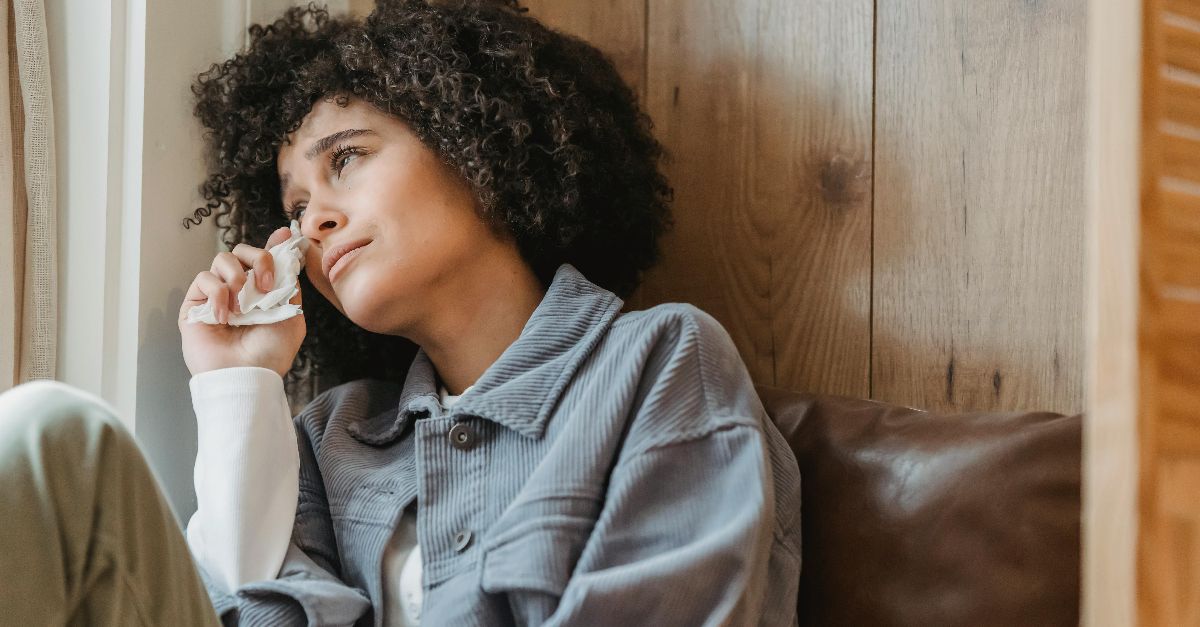 Desperate African American female with curly hair wiping face with tissue while crying from sadness on windowsill and looking away